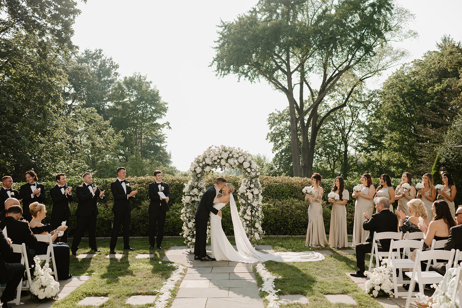 Bride and groom share a first kiss under a lush white rose arch while guests and wedding party applaud.